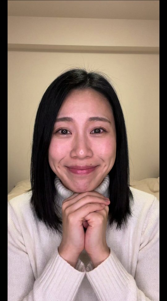Soft pink light, white turtleneck, hands under her chin: she plays the flattering AI.