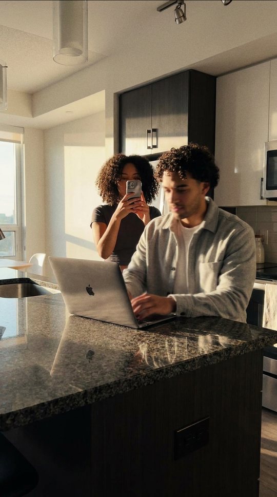 Brother at a kitchen counter with a laptop; sister behind him, phone up.