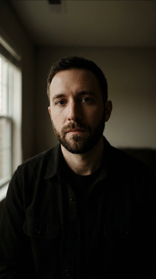 Close-up, low side light, dark room. He looks straight into the lens.