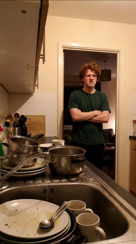 A sink piled with a week of dishes. He stands in the doorway, arms folded.